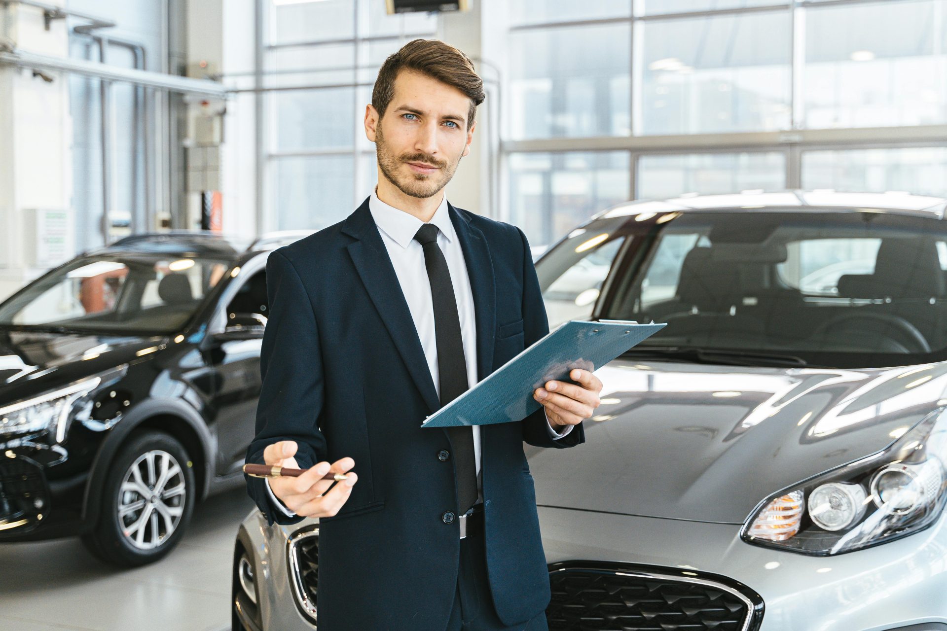 Professional car dealer in business suit holding clipboard in a bright car showroom.